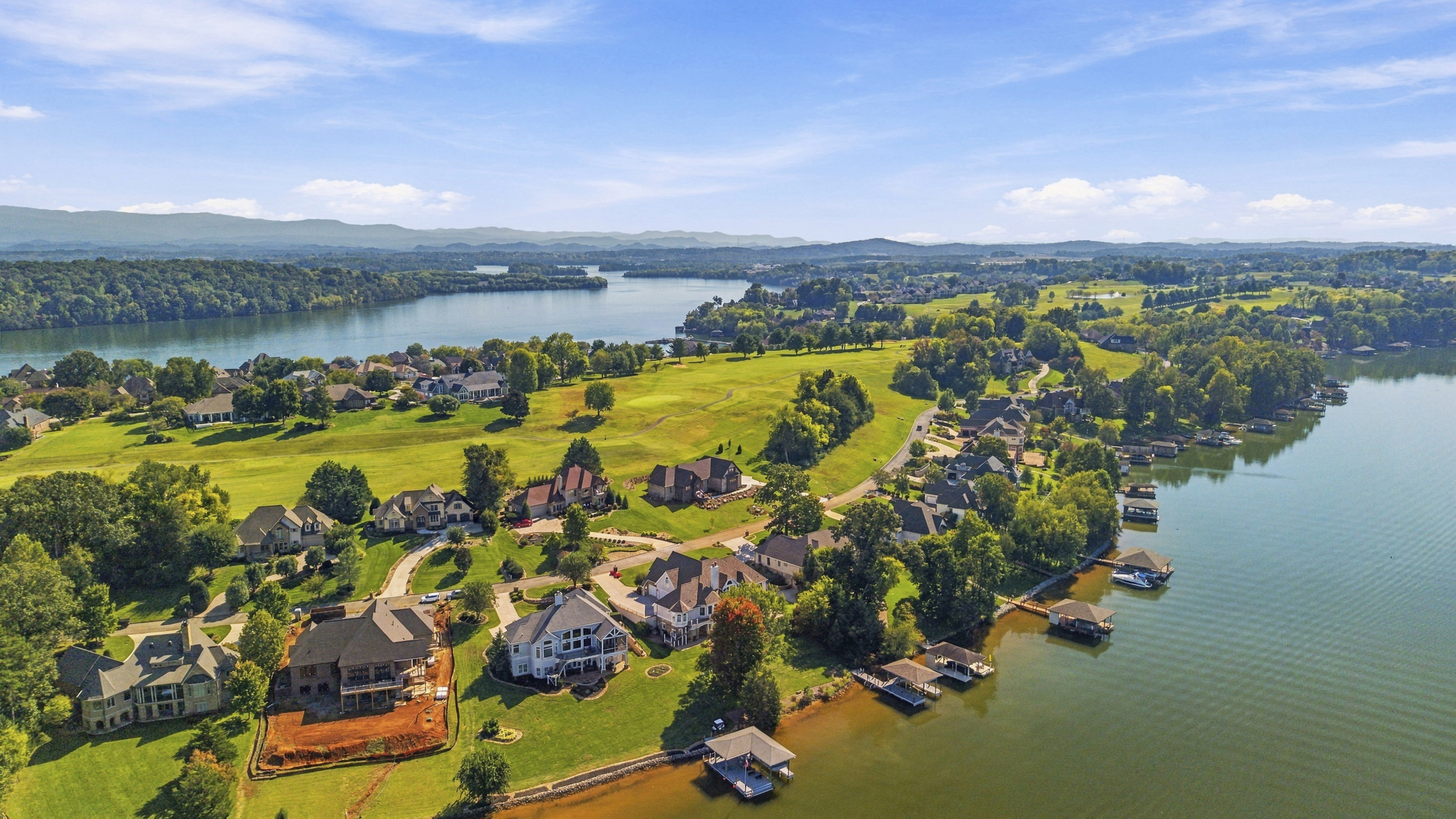 Aerial view of lakeside homes, docks and a green golf course with distant mountain ridges.