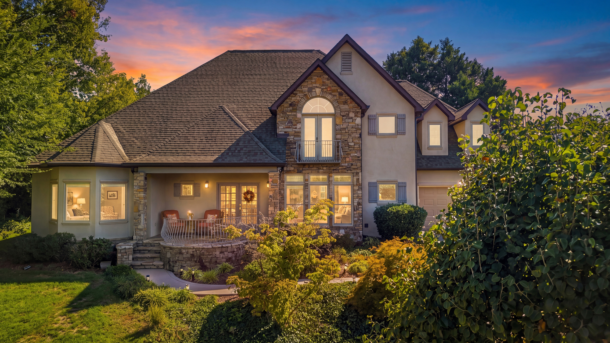 Stone-and-stucco home with glowing windows, landscaped gardens and a pink twilight sky.